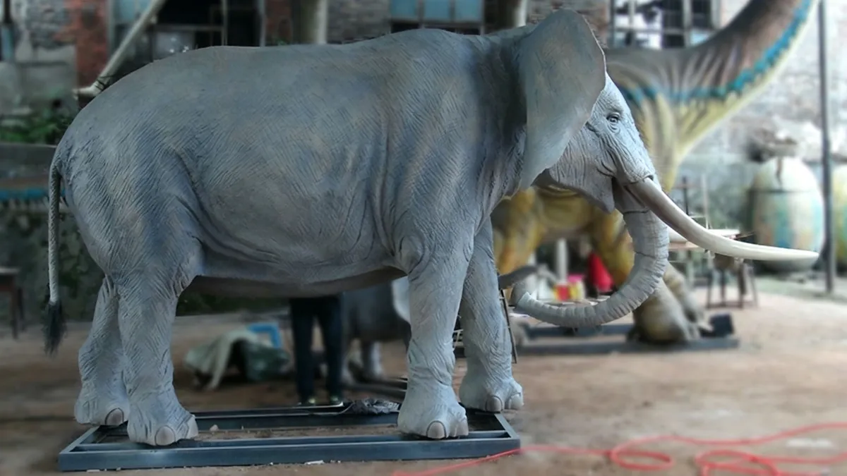 Lifesize animatronic African elephant standing in workshop showing full body profile, realistic grey silicone skin texture and long curved tusks