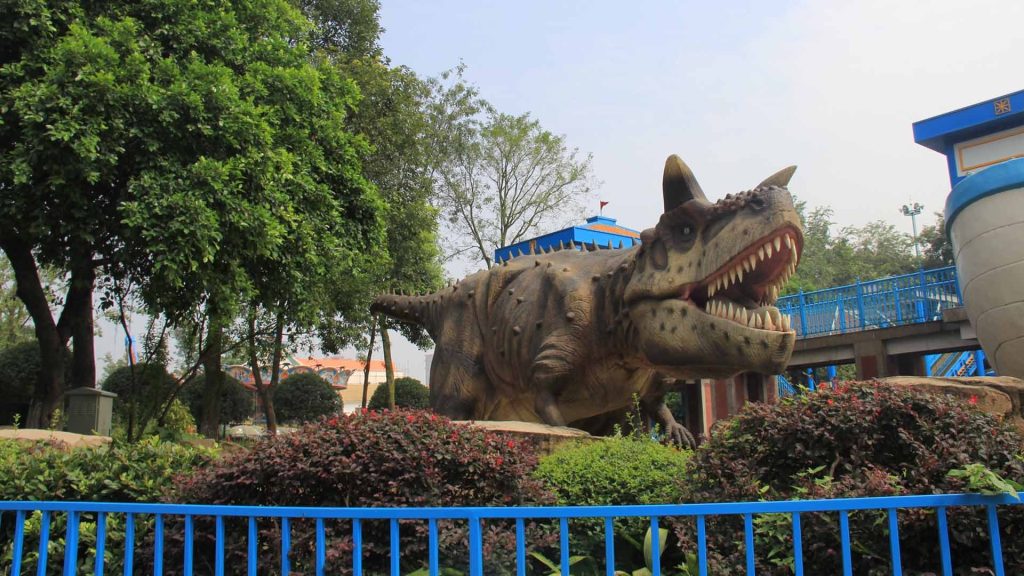 Animatronic Carnotaurus model with horns and exposed teeth sitting among shrubs in a park
