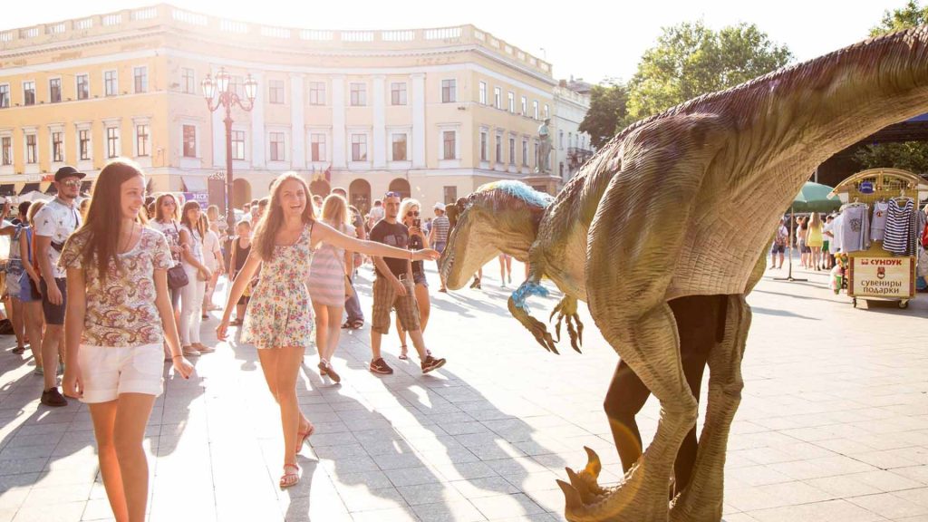 Dinosaur costume performer in a crowd reaching to touch the animatronic T-Rex